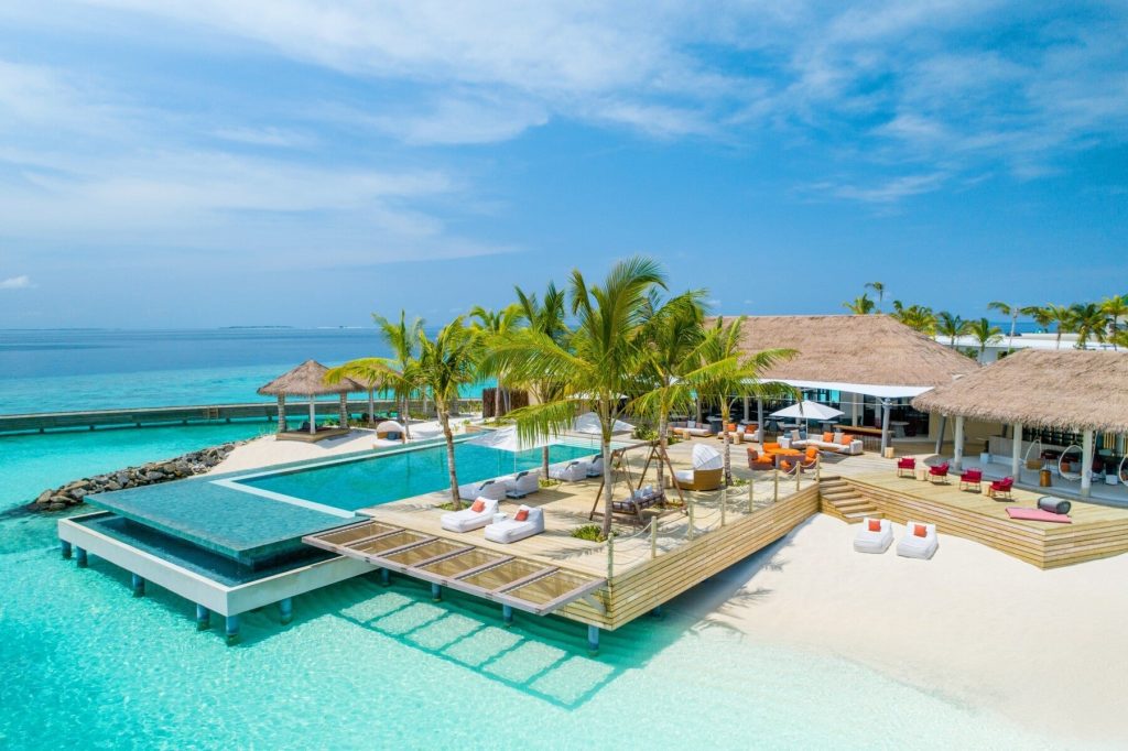 Infinity-Pool mit Holzterrassen und Strohdächern vor türkisfarbenem Meer und grünen Palmen