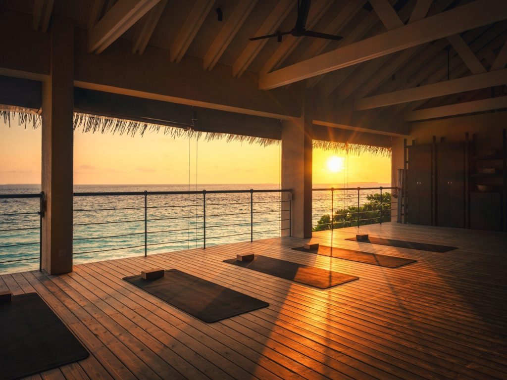 Offener Wellnessraum mit Holzfußboden, Yoga-Matten und Blick auf Meer bei Sonnenuntergang