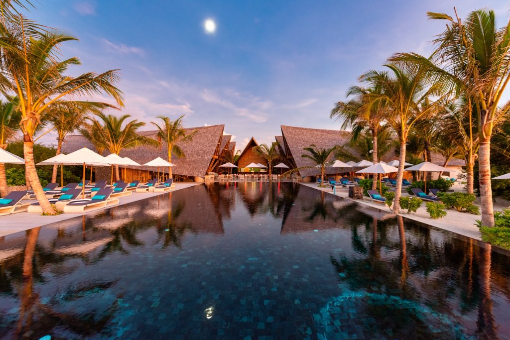 Infinity-Pool umgeben von Palmen und Holzgebäuden mit spitzen Dächern, Sonnenliegen mit blauen Polstern und weißen.