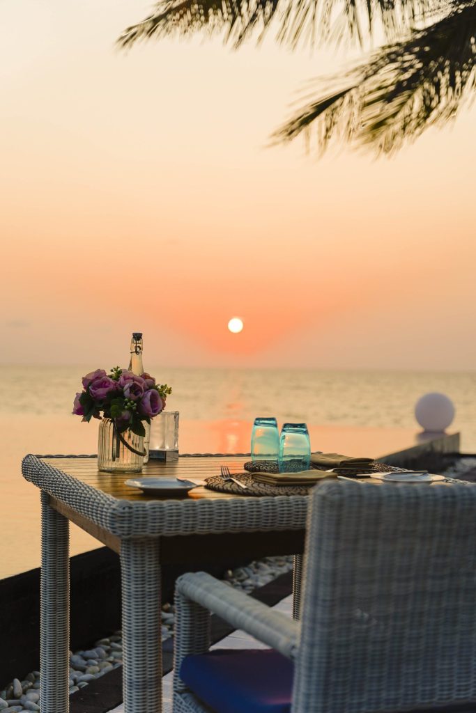 Gedeckter Rattan-Tisch mit blauen Gläsern und lila Blüten unter freiem Himmel mit Meerblick bei Sonnenuntergang.