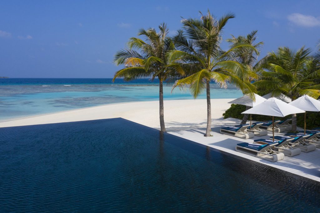 Infinity-Pool mit blauen Liegen und weißen Sonnenschirmen neben weißem Sandstrand und Palmen am Meer