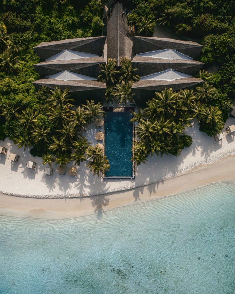 Luftaufnahme des Kuredhivaru Resort and Spa Maldives mit rechteckigem Pool, Palmen, weißen Sandstrand und Hotelgebäuden.