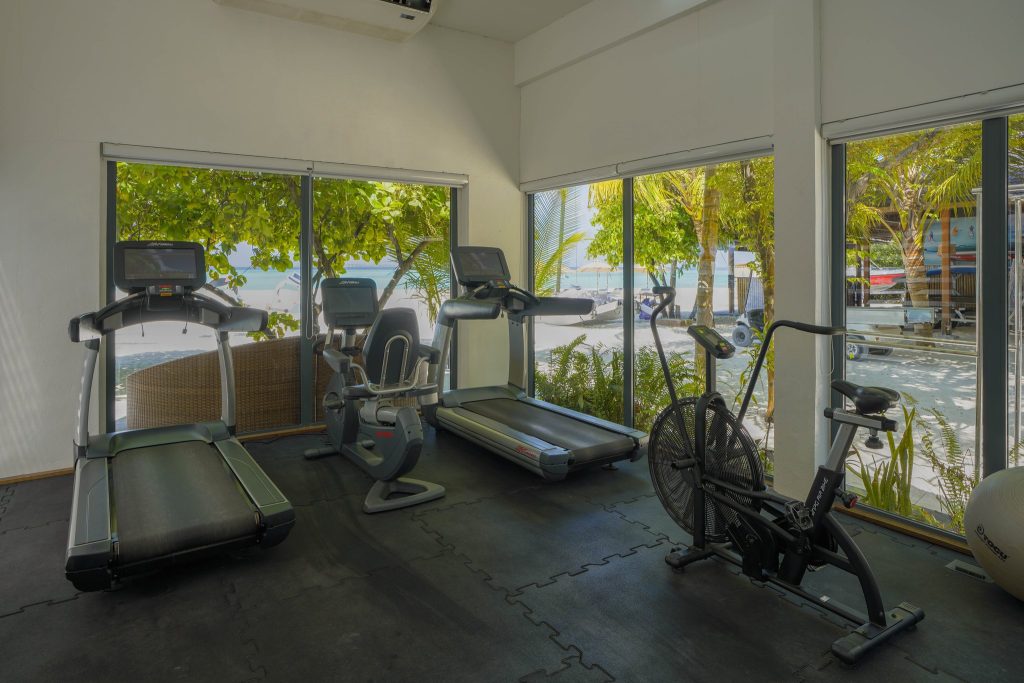 Fitnessraum im Kuredhivaru Resort and Spa Maldives mit Laufbändern, Fahrrad, Crosstrainer und großen Fenstern zum Strand