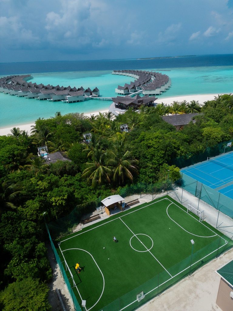 Kunstrasen-Fußballplatz und blauer Tennisplatz im Kuredhivaru Resort and Spa Maldives mit Wasserbungalows und tropischem Grün