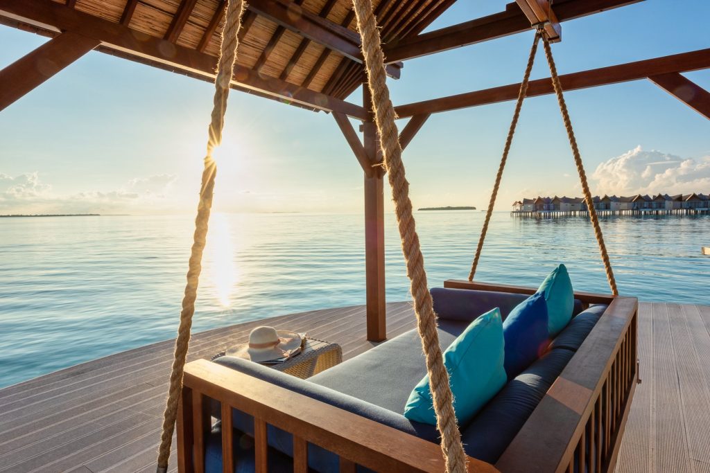 Terrasse mit schwebendem Holzsofa an dicken Seilen über Wasser und Wasserbungalows im Hintergrund