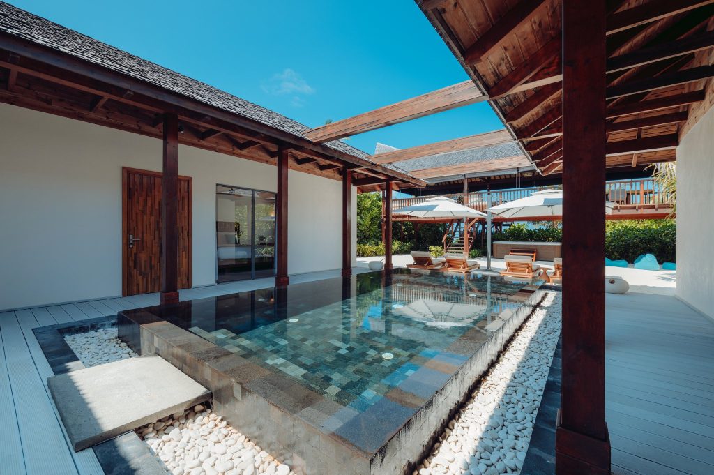 Außenbereich der Four Bedroom Beach Pool Residence mit Pool, Holzdeck, weißen Kieselsteinen und Sonnenliegen in Kuredhivaru.