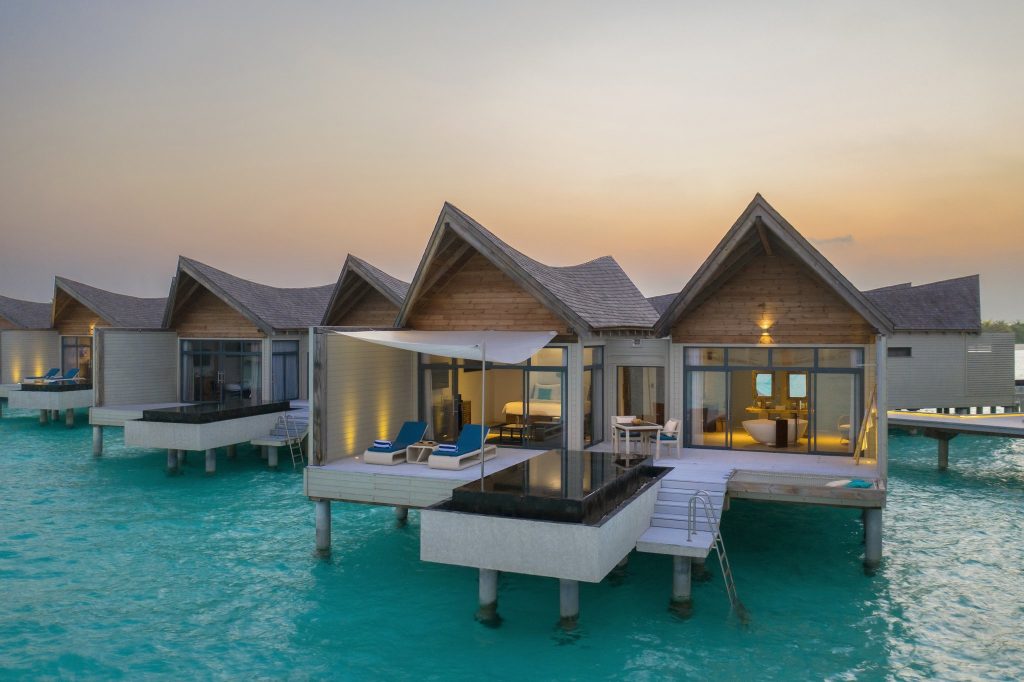 Mehrere Wasser-Villen auf Stelzen mit Holz- und Glaselementen, spitz zulaufenden Dächern und Pools über türkisblauem Meer.