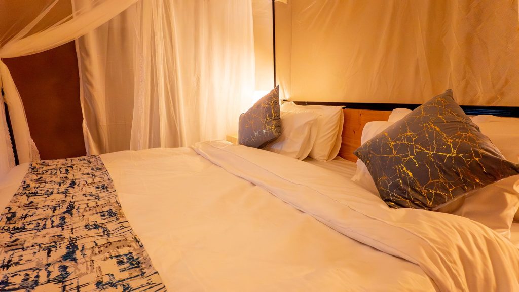 Hotelzimmer im Misako Mara Migration Camp mit weißem Bett, blau-gold gemusterten Kissen und hellen Stoffvorhängen