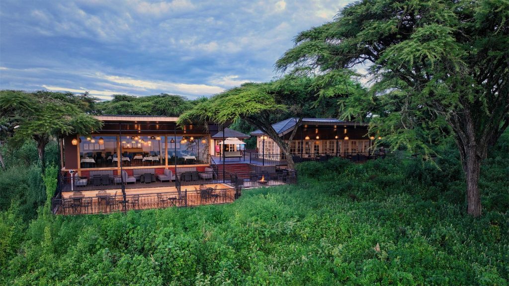 Ngorongoro Lions Paw Außenbereich mit großen Glasfronten Modernes Hotelgebäude mit großen Glasfronten, Holz- und Metallelementen umgeben von grüner Landschaft in Tansania