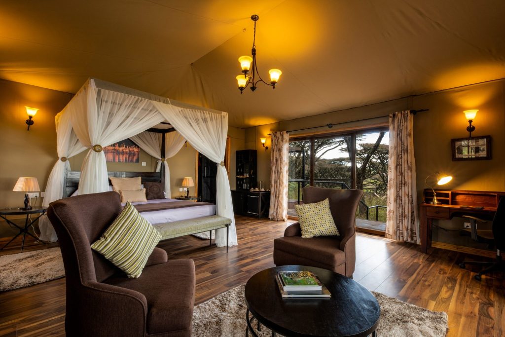 Honeymoon Suite im Ngorongoro Lions Paw mit großem Himmelbett, Holzboden, braunen Sesseln und Schreibtisch mit Fensterblick