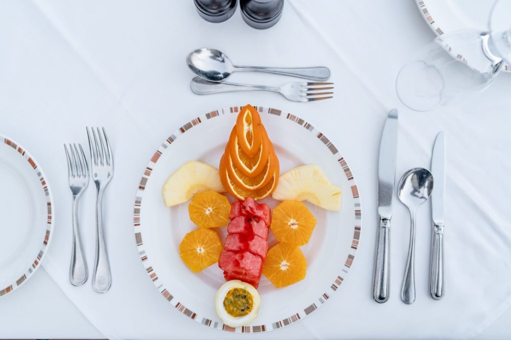 Kunstvoll arrangierter Obstteller mit Orangen, Ananas, Wassermelone und Passionsfrucht auf weißem Tischläufer.