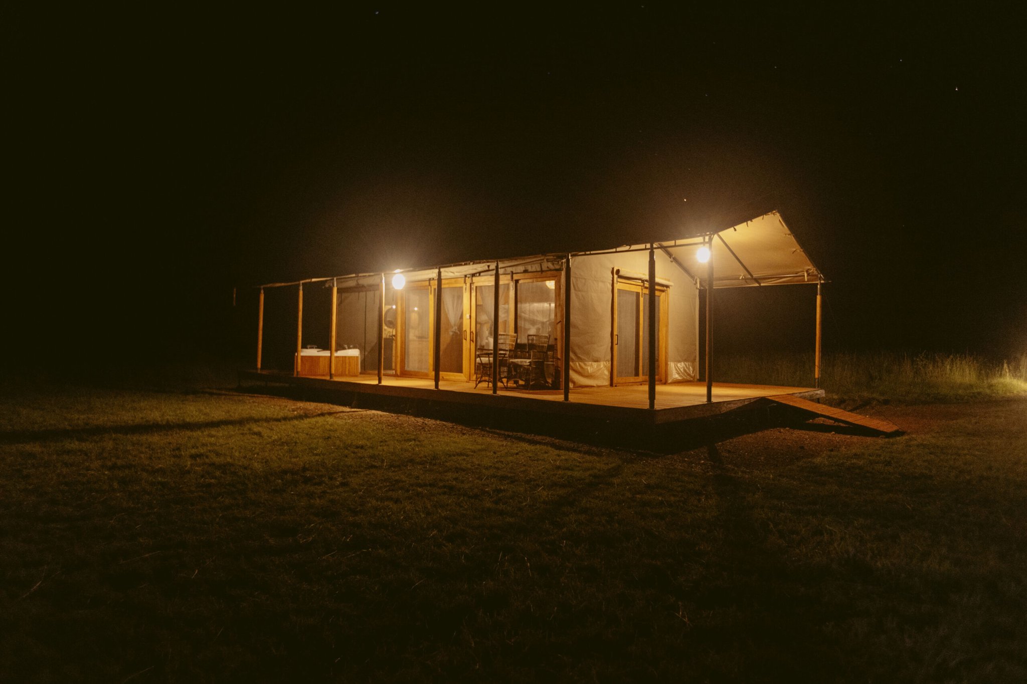 Glamping-Struktur aus Holz, Metall und Stoff mit großen Glasschiebetüren und warmem Licht auf der Veranda