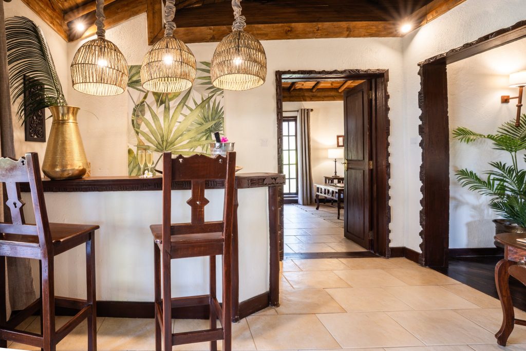 Barbereich im The Palms Zanzibar mit dunklem Holz, zwei Holzstühlen, rattanartigen Hängelampen und Palmenbild an Wand