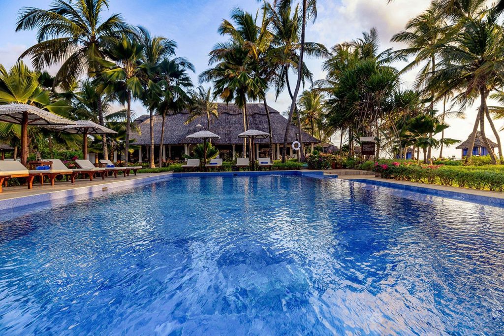 Hotelpool mit klarem blauem Wasser, Holzliegen mit beigen Auflagen und Strohdach-Gebäude im Hintergrund