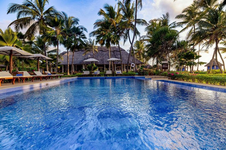 Hotelpool mit klarem blauem Wasser, Holzliegen mit beigen Auflagen und Strohdach-Gebäude im Hintergrund