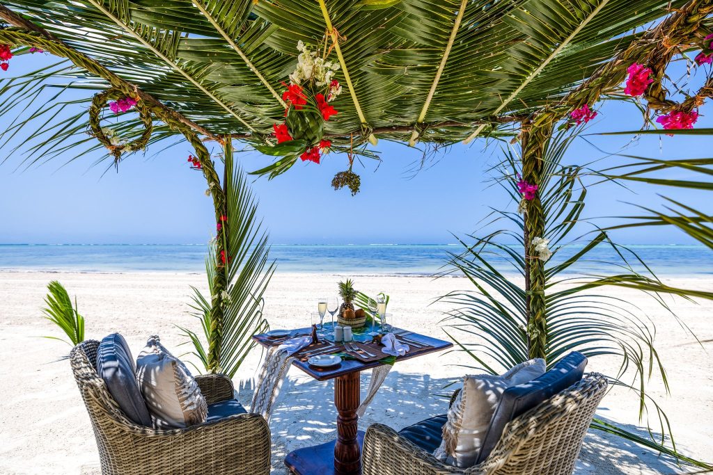 Zwei Rattanstühle mit grauen Kissen an Holztisch mit weißem Tischläufer unter Palmblätter-Pergola am Strand