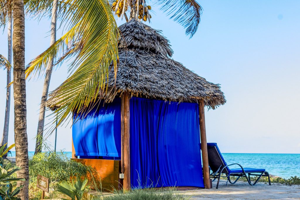 Strandhütte mit blauem Strohdach und blauen Vorhängen, zwei blaue Liegestühle auf Sand vor Palmen und Meer