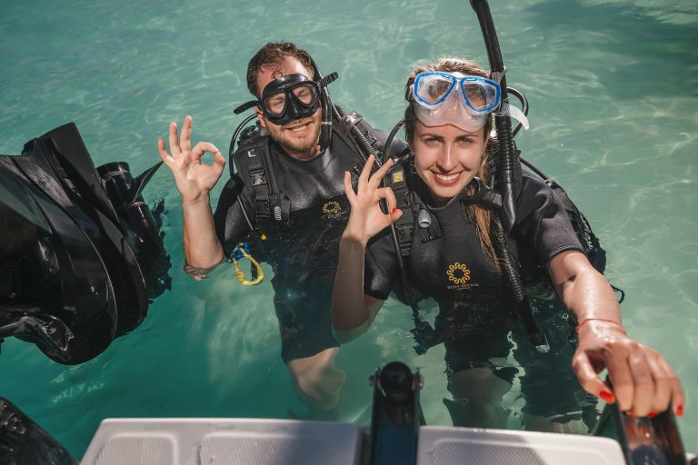 Zwei Personen in Taucheranzügen mit Taucherbrillen und Mundstücken stehen im klaren Wasser neben einem Boot