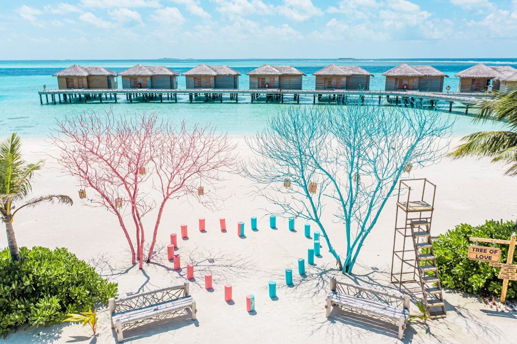 Strand mit zwei bunt bemalten Bäumen in Rosa und Blau, weißer Sand und Überwasserbungalows im Meer auf den