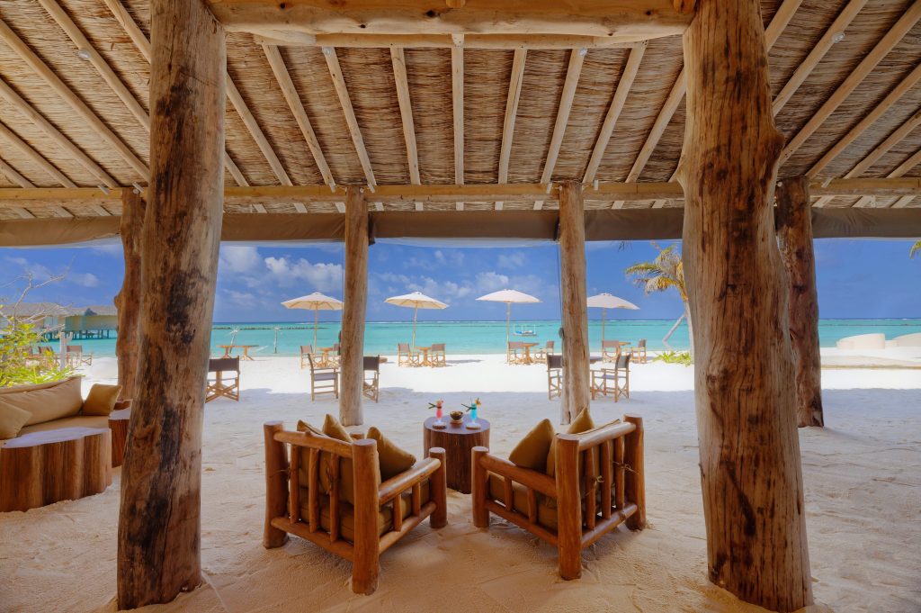 Offener Lounge-Bereich der Cheers Bar mit Holzsesseln, beigen Kissen, Sandboden und Blick auf Strand und Meer