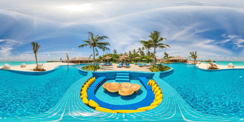 Großer türkisfarbener Pool mit blauen Fliesen und gelben Polstern, weißer Sandstrand und Palmen im Hintergrund