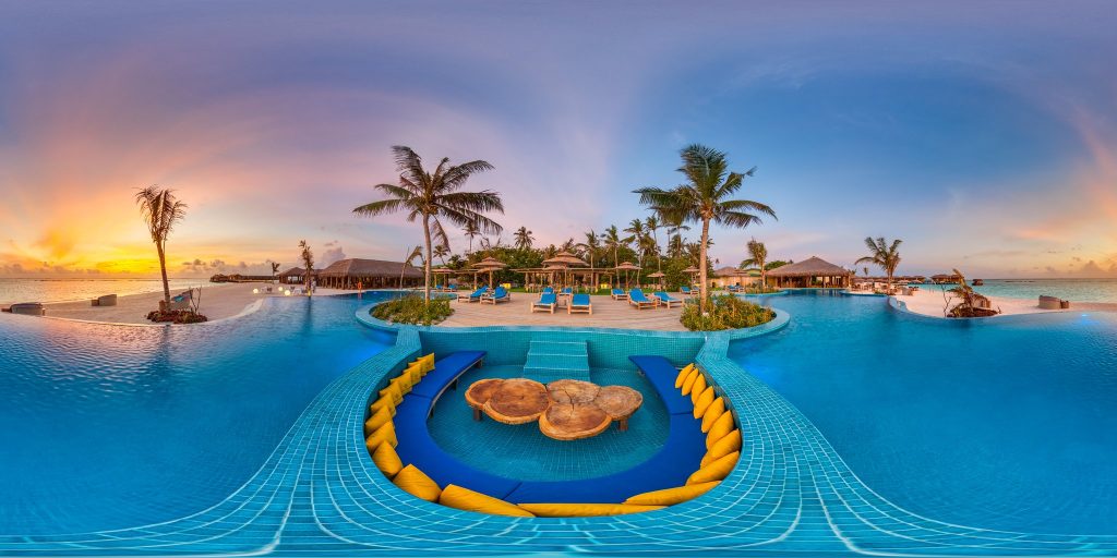 Großer geschwungener Pool mit hellblauen Fliesen, Sitzecke mit blauen Kissen, Palmen und Liegestühle auf Holzterrasse.