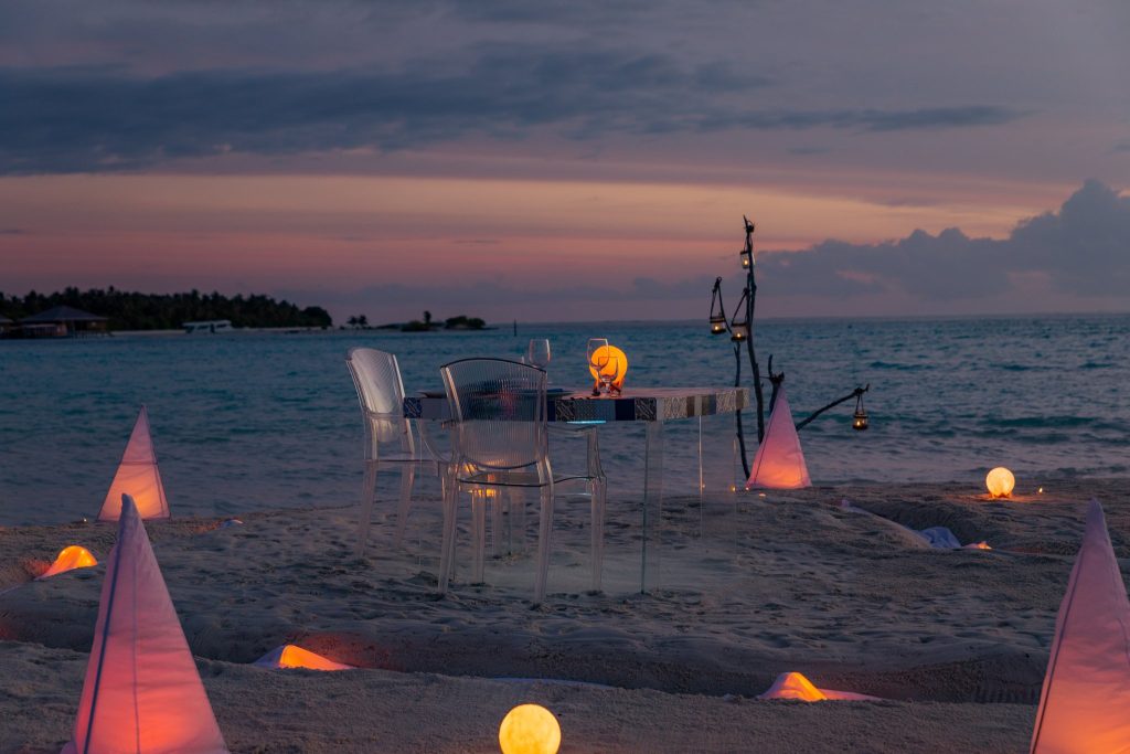 You & Me Maldives restaurant mit Glas-Tisch und durchsichtigen Stühlen am Strand bei Dämmerung und Lampenlicht