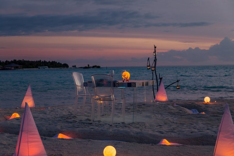 You & Me Maldives restaurant mit Glas-Tisch und durchsichtigen Stühlen am Strand bei Dämmerung und Lampenlicht