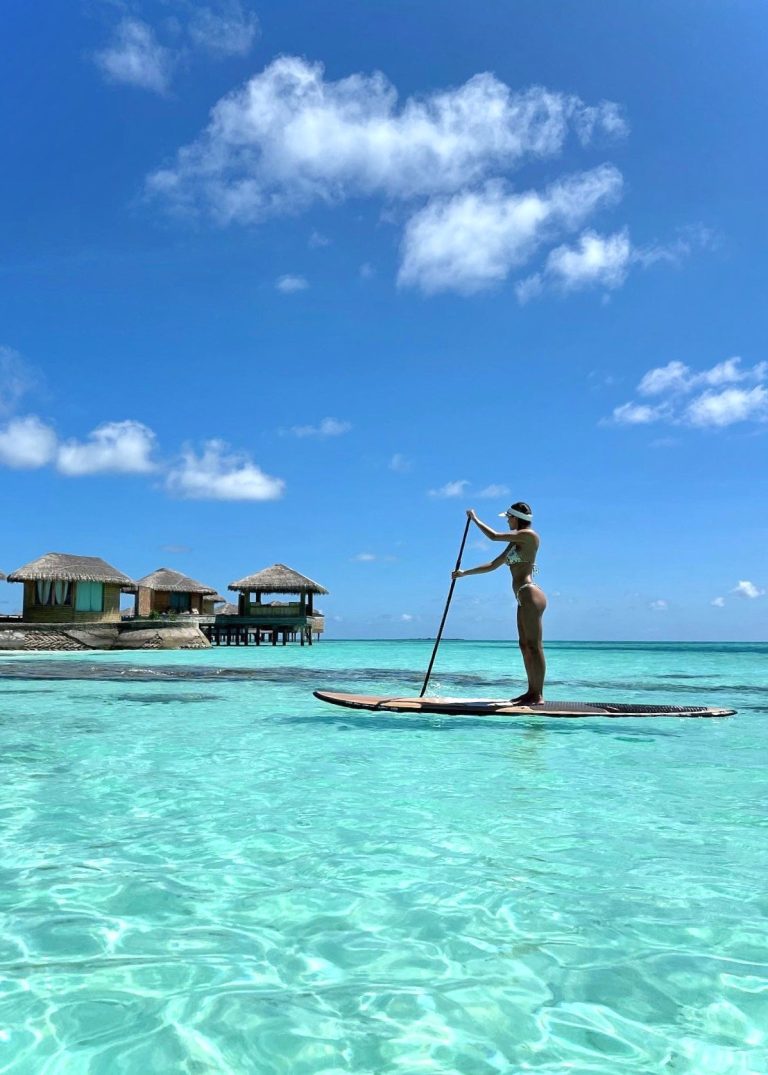 Person paddelt auf Stand-up-Paddleboard vor Bungalows mit Strohdächern über Wasser auf den