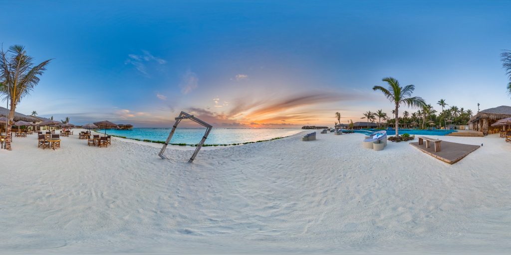 Weitläufige Strandlandschaft bei You & Me Maldives mit Holzmöbeln, Pool und Palmen unter Sonnenuntergangshimmel