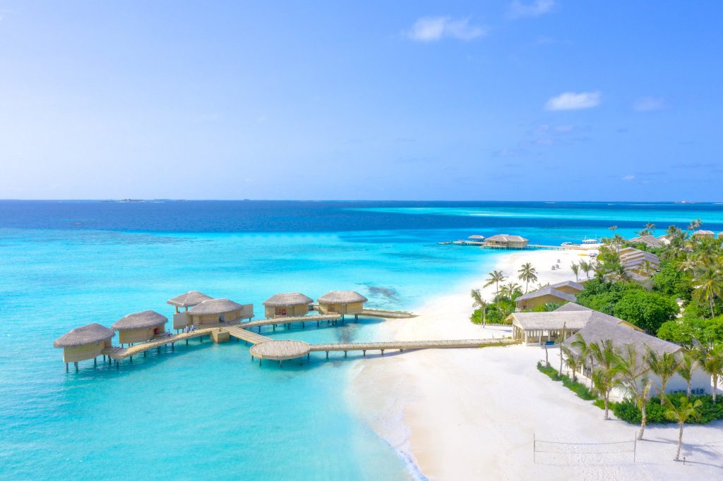 Luftaufnahme von Bungalows auf Stelzen mit Strohdächern über türkisfarbenem Wasser und weißem Sandstrand mit Palmen