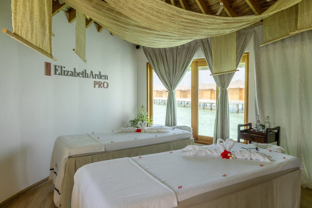 Spa-Raum mit zwei Massageliegen, weißen Handtüchern, roten Blüten, Holzdecke und Blick auf blaue Gewässer.