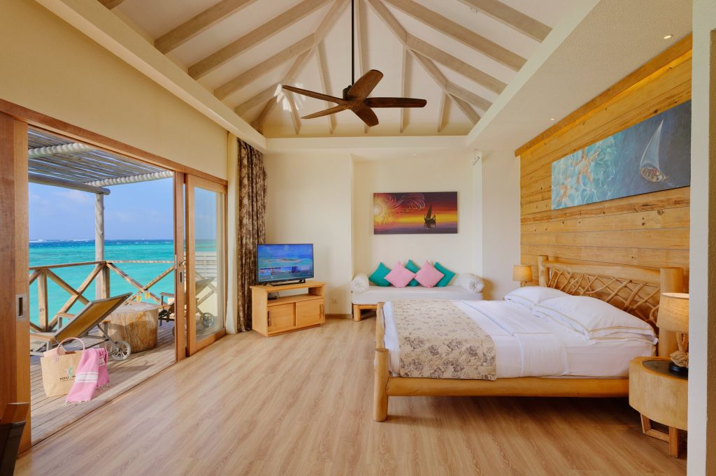 Zimmer der Aqua Suite im Hotel You & Me Maldives mit Bett, Holzmöbeln und Meerblick durch Glasschiebetür