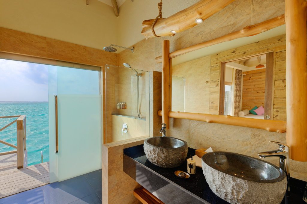 Badezimmer der Lagoon Suite im You & Me Maldives mit Doppelwaschbecken, Stein- und Holzelementen und Blick auf das Meer