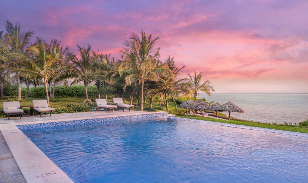 Rechteckiger Pool mit klarem blauem Wasser, beige Sonnenliegen auf Steinterrasse, Palmen und Meer im Hintergrund.