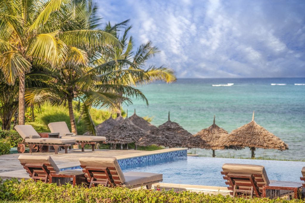 Tropischer Hotelpool mit Holzliegen, beigen Auflagen, Palmen und Strohdächern am Meer in Sansibar