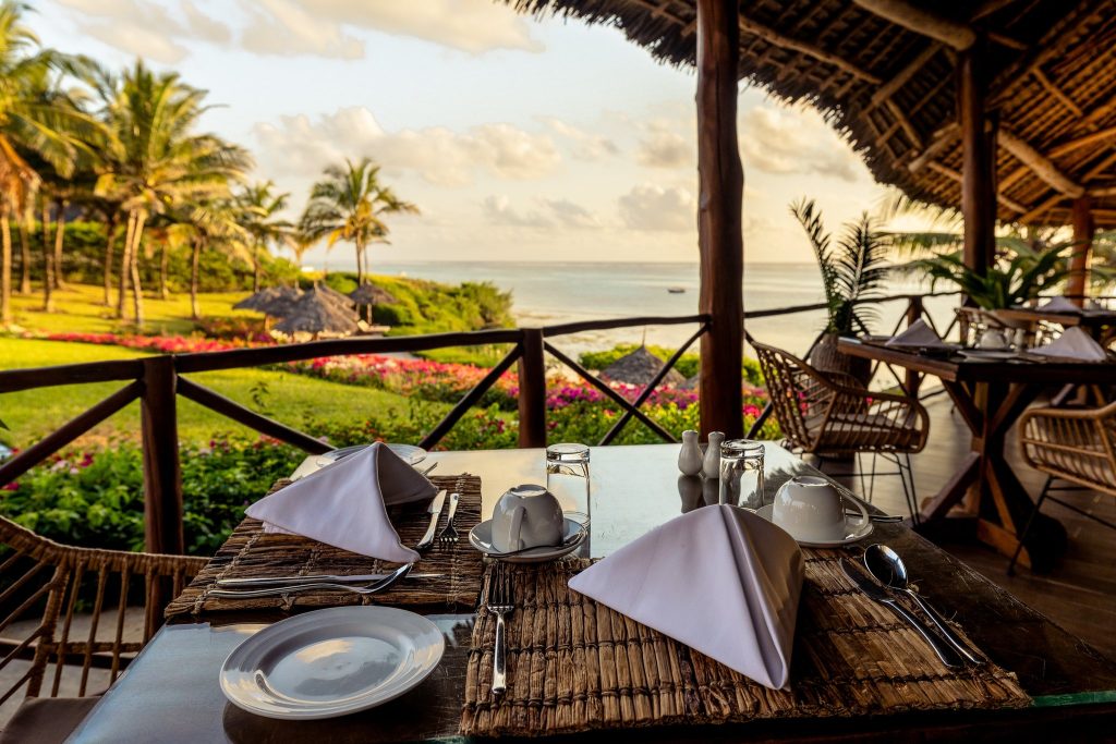 Terrasse des Restaurant Drift Wood im Zawadi Hotel mit gedecktem Tisch, Rattanstühlen und Blick aufs Meer