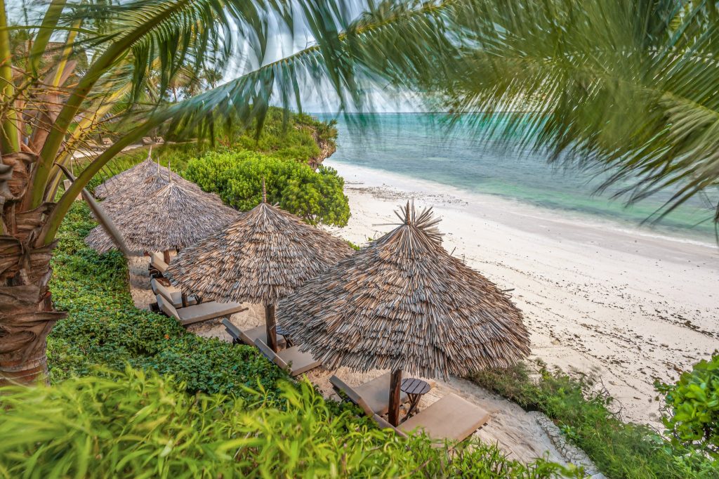 Strandabschnitt mit Liegen unter Palmblatt-Schattendächern und üppiger grüner Vegetation am Zawadi Hotel Sansibar