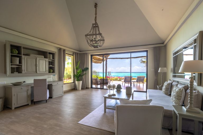 Helles Hotelzimmer mit großem Fenster, grauem Sofa, weißem Couchtisch, Schreibtisch und Blick auf Terrasse und Meer