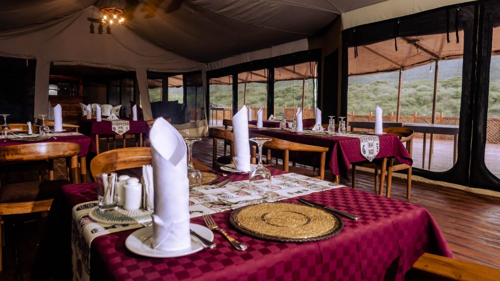 Restaurantbereich im Zelt mit weinroten Tischdecken, geflochtenen Platzdeckchen, Holztischen und großen Fenstern in Tansania