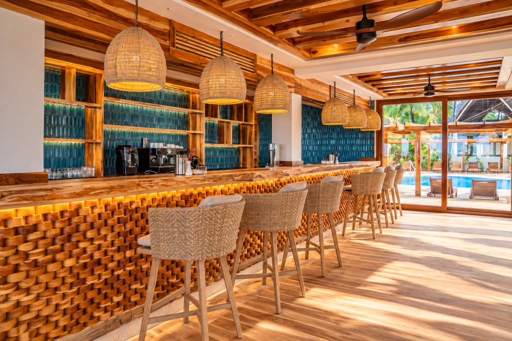 Bar im JAZ Elite Adonia mit hellem Holztheke, geflochtenen Barhockern, türkisfarbener Wand und Blick auf Pool und Palmen