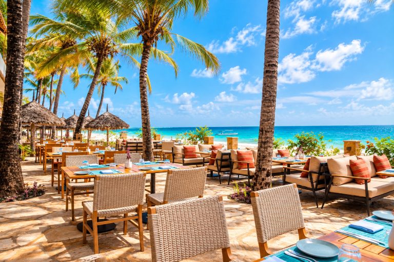 Außenbereich des JAZ Elite Adonia Strandrestaurants mit Holztischen und Blick auf den Ozean und Palmen