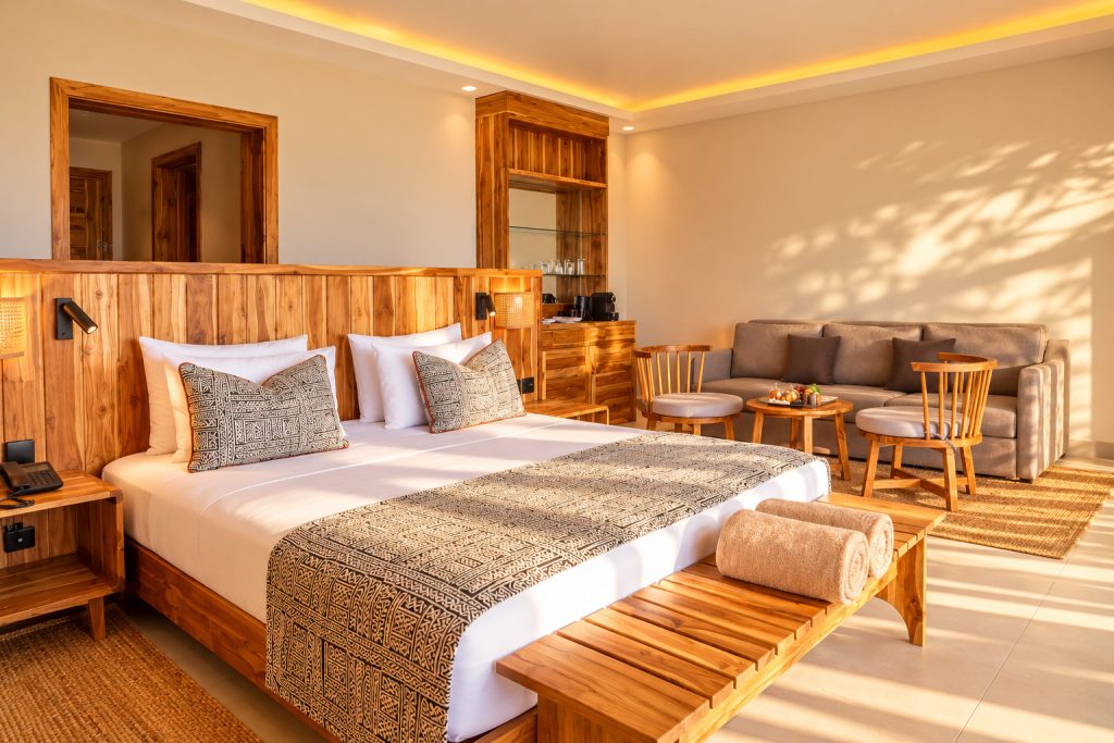 Geräumiges Hotelzimmer mit großem Bett, Holz-Nachttisch, grauer Couch, zwei Holzstühlen und Tageslicht in Sansibar