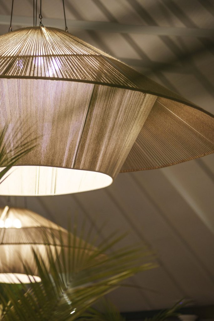 Hängelampe aus Stoffgeflecht in Beige und Gold mit Schatten an hellem Holzdecke, grüne Blätter im Vordergrund