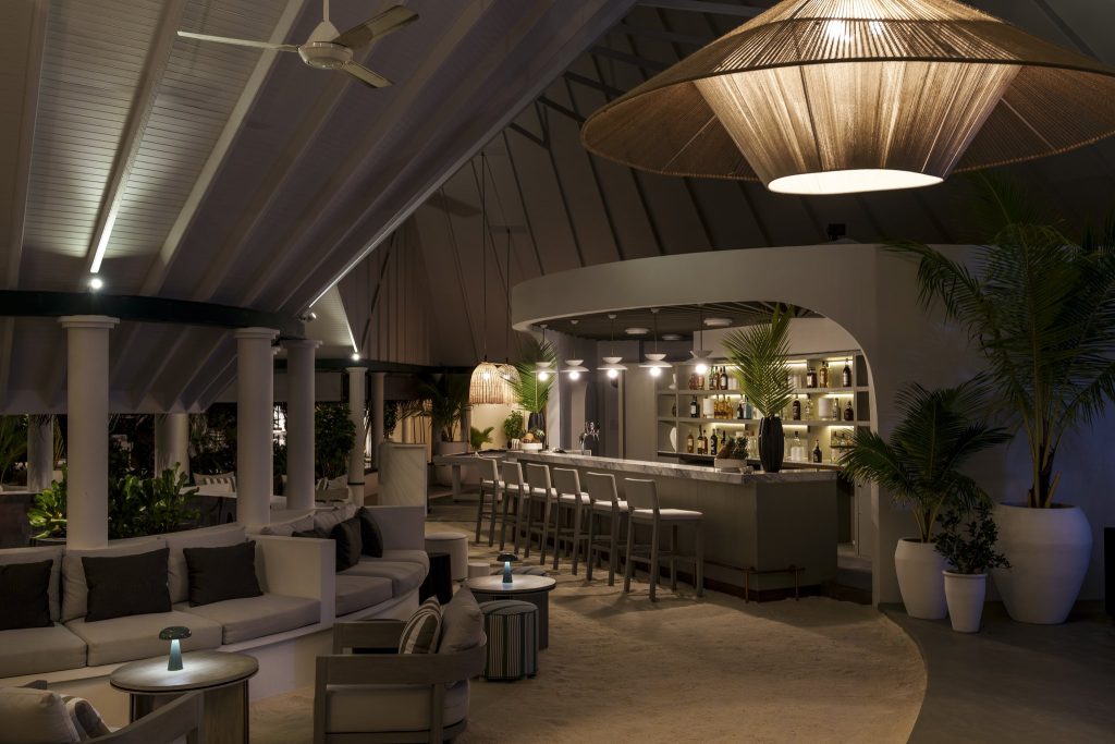 Bar Nautilus mit geschwungenen Sofas, runden Holztischen, Pflanzen und dekorativer Deckenbeleuchtung im Sun Siyam Vilu Reef