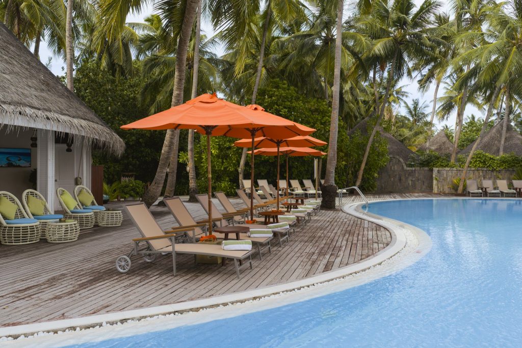 Poolbereich im Sun Siyam Vilu Reef mit Holzdeck, Liegestühlen, orangefarbenen Sonnenschirmen und Palmen in den