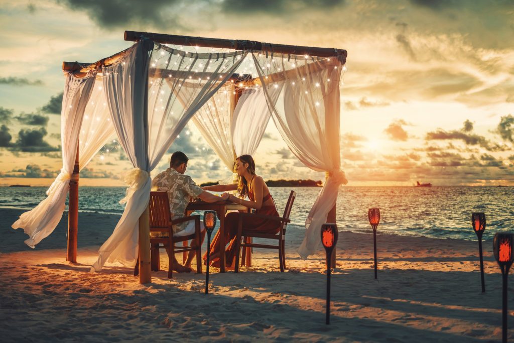 Romantisches Dinner-Setup im Pavillon mit weißen Vorhängen am Strand bei Sonnenuntergang im Sun Siyam Vilu Reef
