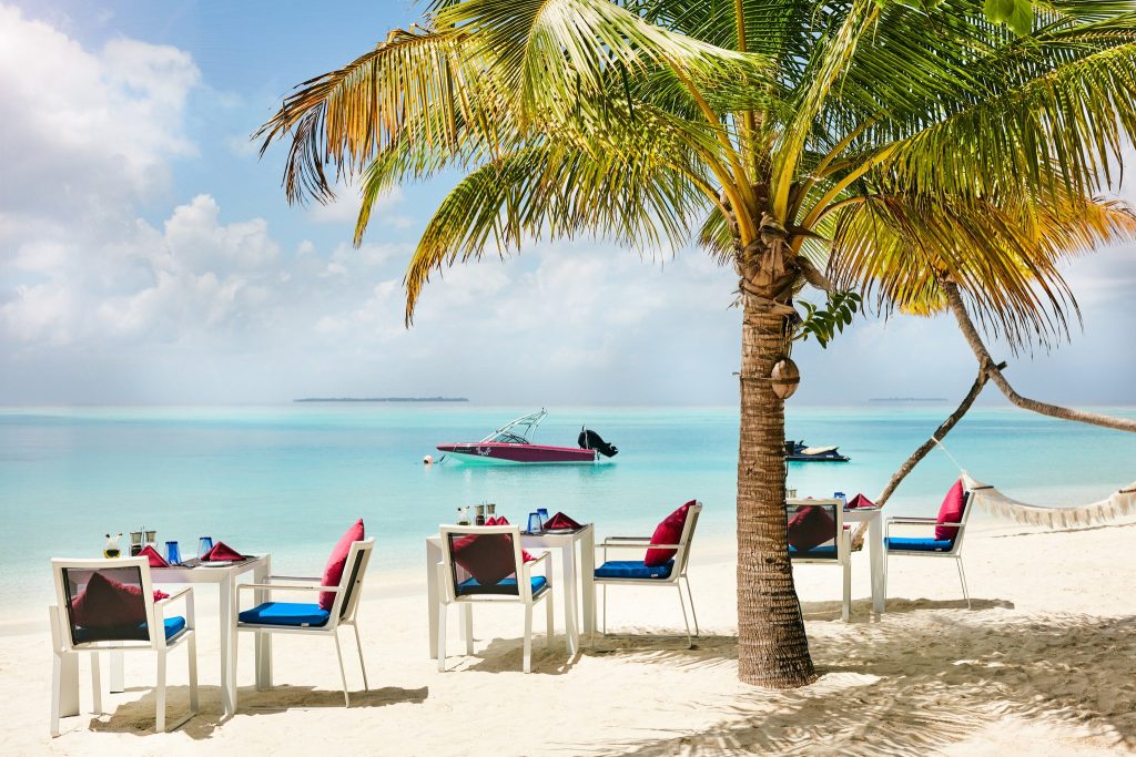 Weiß gestrichene Tische und Stühle mit roten und blauen Kissen auf hellem Sand, Palme rechts.
