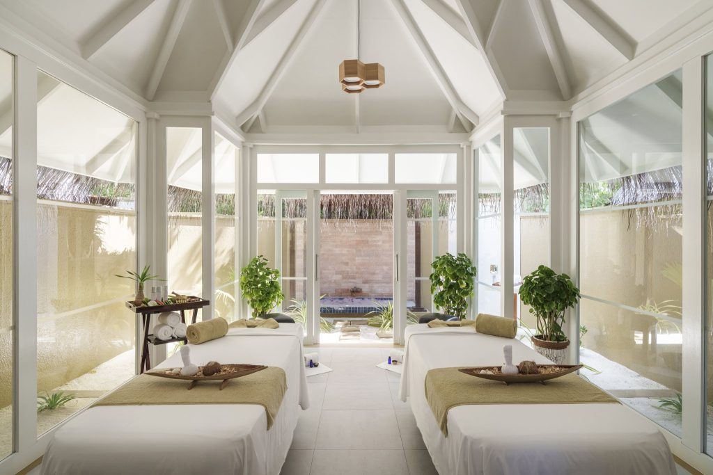 Hell beleuchteter Wellness Spa Raum mit zwei Massagebänken, großen Fenstern, grünen Pflanzen und Holzdecke im Sun Siyam Vilu.