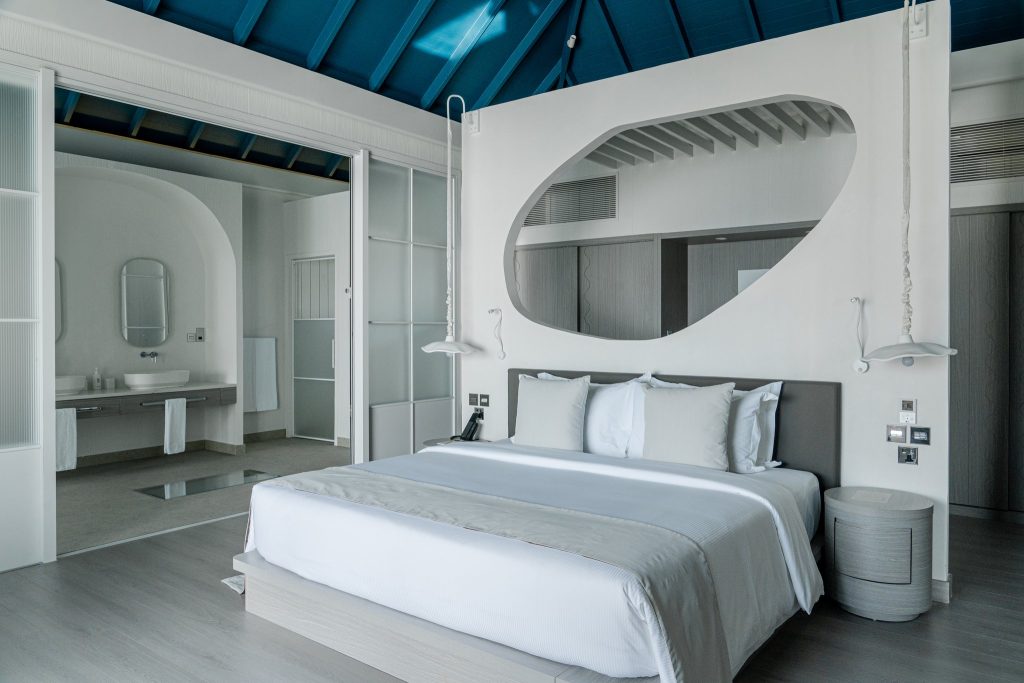 Modernes Hotelzimmer 2BR Ocean Signature Villa mit großem Bett, ovalem Spiegel und angrenzendem Badezimmer im Sun Siyam Vilu.